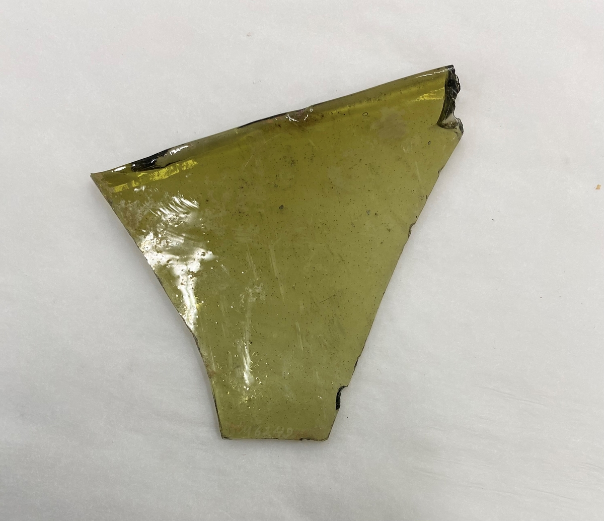 Del av ett fönsterglas med grön glasmassa. Ena sidan är mjukt rundad och de övriga är avslagna. Vid den rundade kanten är en yta med tunna diagonala roströda streck. Möjligen spår efter att den varma cylinderglasrutan planats ut.