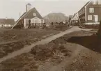 Fotografi från expedition till Grönland. Motiv av by med berg i bakgrunden. På bilden syns ett antal hus varav ett är en kyrka. Bilden är tagen i Holstenborg/Sisimiut som numera är huvudort i Qeqqata kommun. Holstenborg/Sisimiut har varit bebott i ungefär 4500 år.