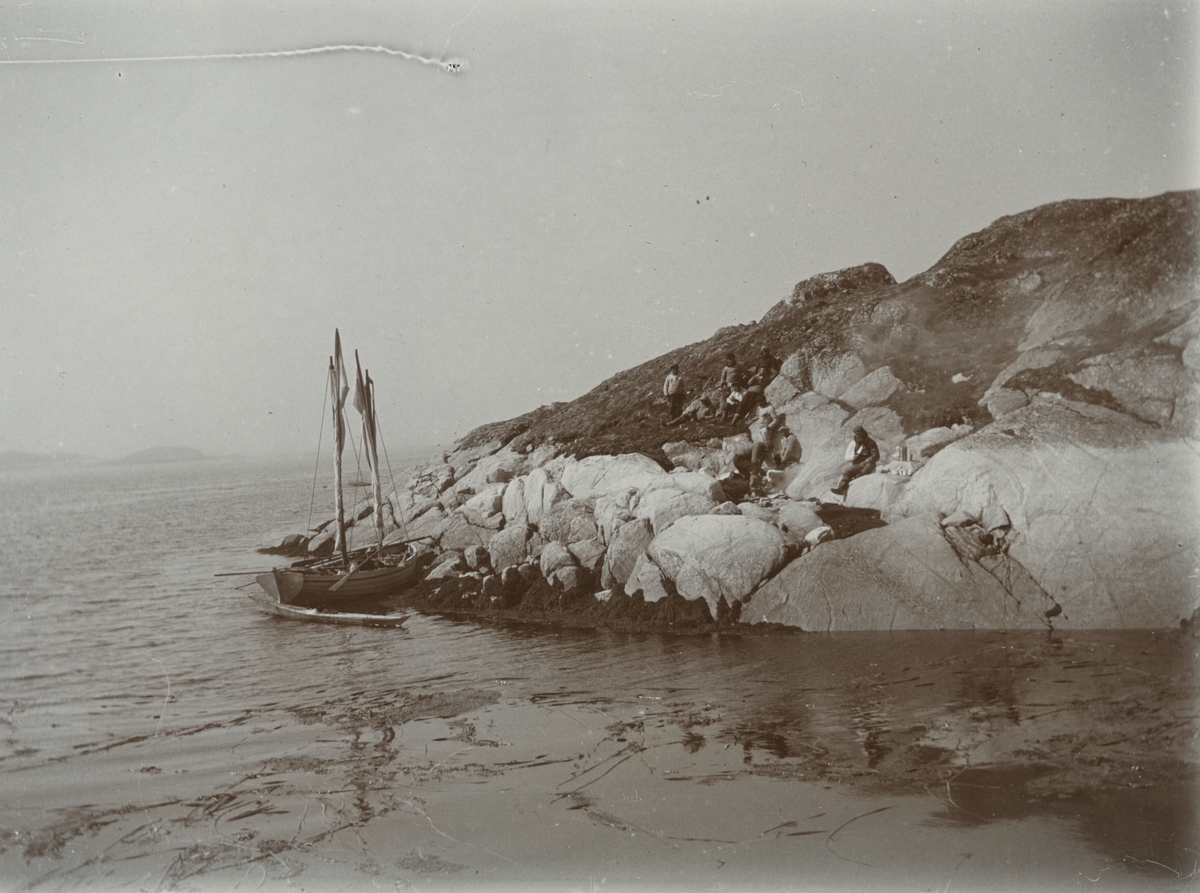 Fotografi från expedition till Grönland. Vy över stor klippa vid havet där några fiskare vilar sig. Deras båt ligger förtöjd strax nedanför klippen.