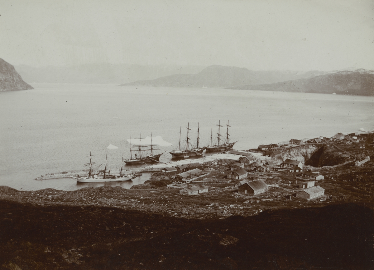 Fotografi från expedition till Grönland. Motiv av liten by vid kustlinjen. I havet vid stranden ligger fyra fiskebåtar.