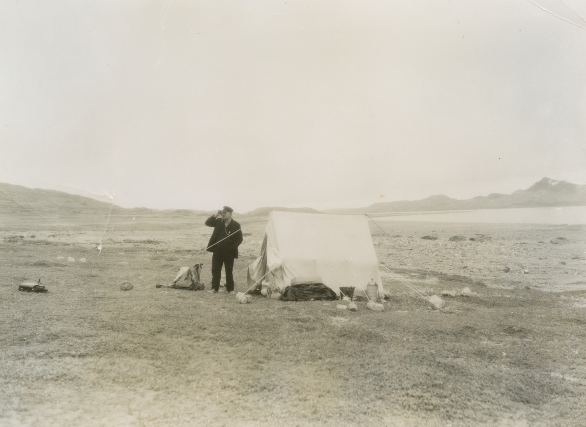 Fotografi från expedition till Grönland. Motiv av Otto Nordenskjöld som står utanför ett vitt tält och tittar i en kikare. Tältet är uppställt mitt i ett kargt och vidsträckt landskap.
