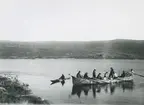 Fotografi från expedition till Grönland. Motiv av nio personer i en båt nära stranden. Till vänster om dem är en person i en kanot. I bakgrunden ses vidsträckta berg.