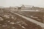 Fotografi från expedition till Grönland. Motiv av ett par större hus i ett bergslandskap. Bredvid husen står en falggstång med flagga i toppen. Det går en väg upp till husen.