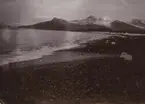 Fotografi från första svenska Antarktisexpeditionen 1901-1904. Vy över stenig strand, hav och snötäckta berg.