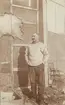 Fotografi från första svenska Antarktisexpeditionen 1901-1904. Motiv av mustaschprydd expeditionsdeltagare som står framför hus.