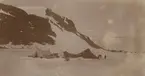 Fotografi från första svenska Antarktisexpeditionen 1901-1904. Motiv av tält och packning i snön nedanför ett berg.