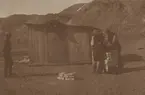 Fotografi från första svenska Antarktisexpeditionen 1901-1904. Motiv av tre expeditionsdeltagare vid träskjul nedanför berg.