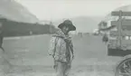 Fotografi från expedition till Peru 1920. Motiv av ung aman med hatt som står på grusväg i by.