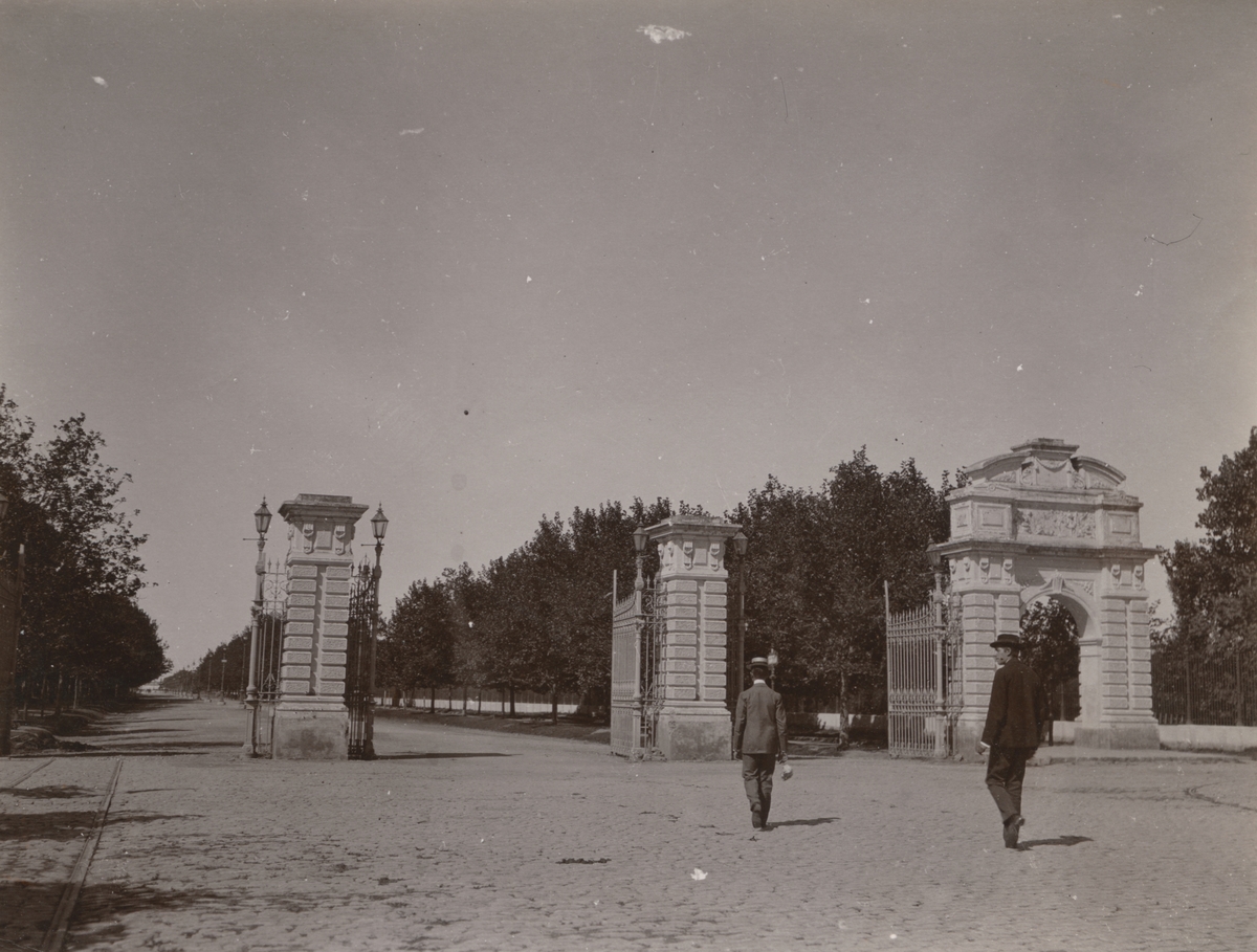 Fotografi från expedition till Peru 1920. Motiv av två män gående mot portar som leder till trädallé.