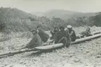 Fotografi från expedition till Peru 1920. Motiv av en liten grupp människor som sitter på en träflotte på en stenig strand i djungeln.