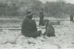 Fotografi från expedition till Peru 1920. Motiv av man och pojke som sitter på stenig strand i djungeln.