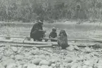 Fotografi från expedition till Peru 1920. Motiv av man och två barn som sitter vid flotte på stenig strand vid flod.