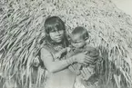 Fotografi från expedition till Peru 1920. Porträtt av kvinna med barn i famnen framför hydda med halmtak.