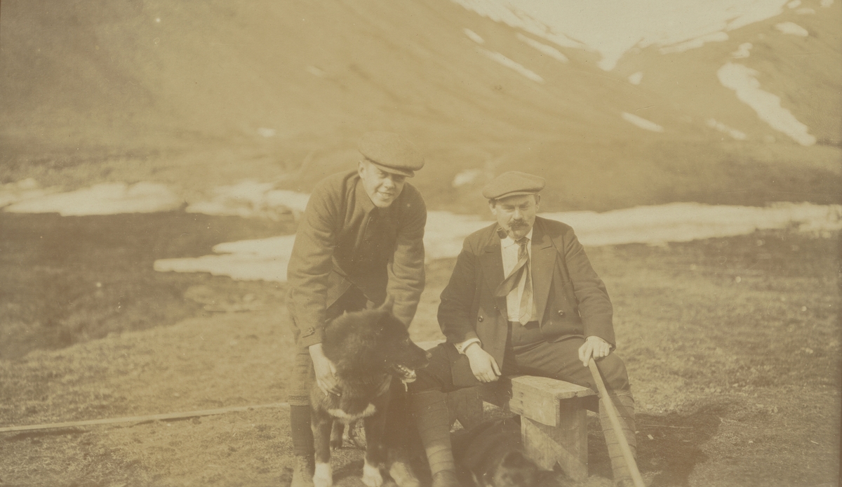 Fotografi från expedition till Spetsbergen. Motiv av två män och en hund sittande nedanför ett berg.