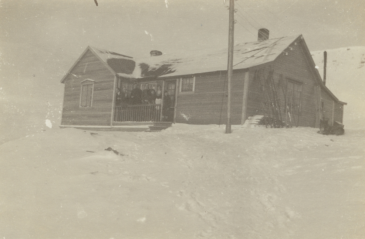 Fotografi från expedition till Spetsbergen. Motiv av en grupp människor som står på en veranda till ett trähus beläget mitt i ett snölandskap.