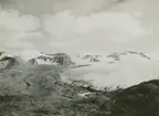 Fotografi från expedition till Östgrönland 1929. Dansk-svensk expedition under ledning av Lauge Koch. Svenska deltagare: Oscar Kulling och Helge Backlund. Vy över glaciär, snötäckt bergslandskap och molnprydd himmel.