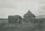 Fotografi från expedition till Peru 1920. Motiv av två stenhyddor med halmtak. Utanför ena hyddan finns en vuxen och två barn och en hund.