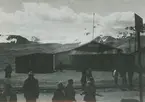 Fotografi från expedition till Peru 1920. Motiv av ett antal människor som står vid ett järnvägsspår utanför en pub. I bakgrunden syns ett snötäckt berg.