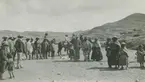 Fotografi från expedition till Peru 1920. Motiv av en grupp hattprydda människor i ett bergigt ökenlandskap. På bilden syns även en häst och hundar.