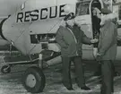 Fotografi från låda märkt Bernt Balchen. Balchen var norsk-amerikansk flygare, polarforskare och militär. Motiv av två räddningsarbetare som samtalar bredvid flygplan. På flygplanet står 