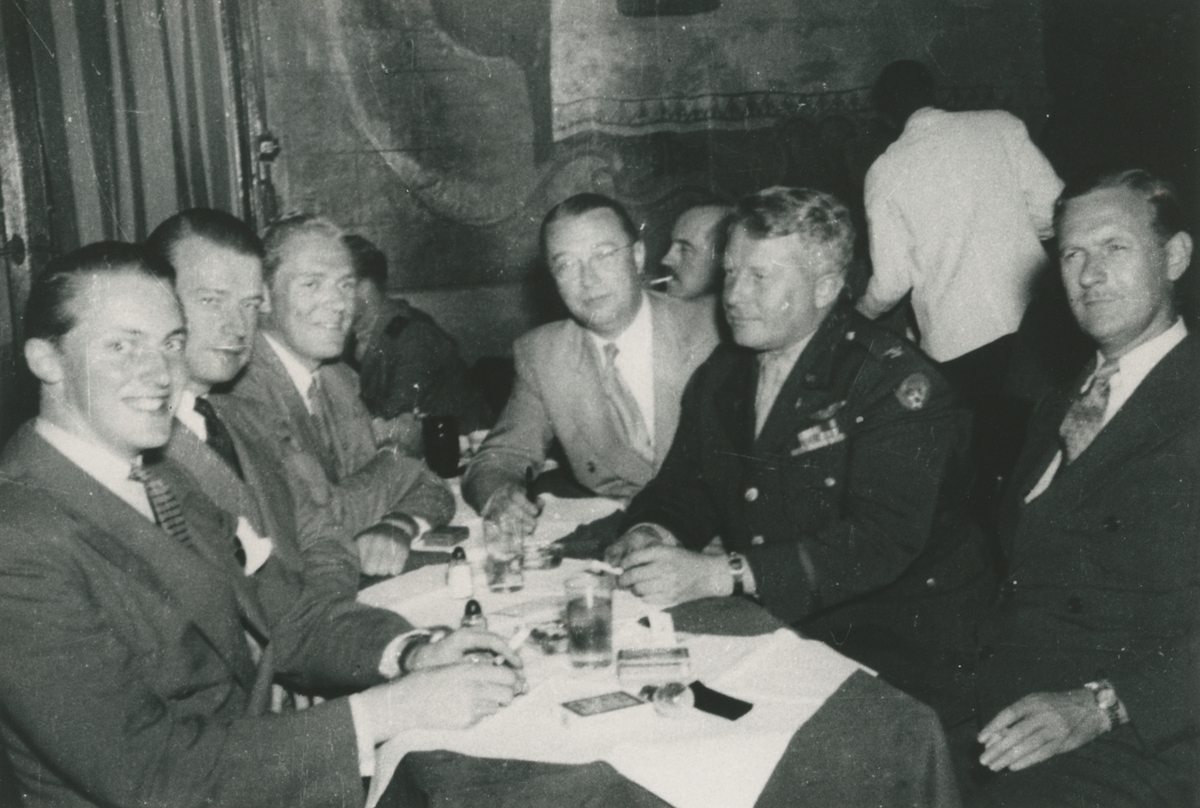 Fotografi från låda märkt Bernt Balchen. Balchen var norsk-amerikansk flygare, polarforskare och militär. Motiv av Balchen och en grupp män som sitter vid bord på restaurang.