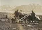 Fotografi från expedition till Sveagruvan. Motiv av fyra expeditionsdeltagare vid lägerplats på stenig strand nedanför berg.