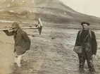 Fotografi från expedition till Sveagruvan. Motiv av två män som vadar genom vatten nedanför berg. En av männen bär ett stativ.