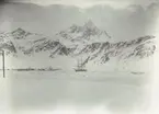 Bild från expedition i Antarktisområdet. Vy över istäckt landskap och berg. Nedanför berget syns en liten hamn och fartyg.