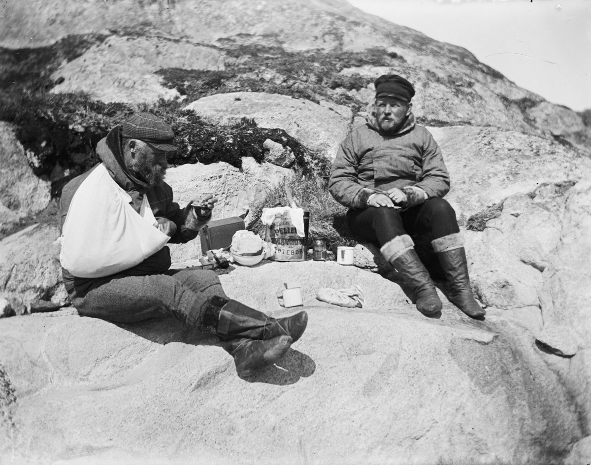 Fotografi från Grönland. Motiv av två män som äter middag på en klippa. Mannen till höger kan vara Otto Nordenskjöld.