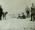 Fotografi från expedition till Spetsbergen. Motiv av fem män och båt på strand.