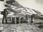 Fotografi från svenska undsättningsexpeditionen 1928. Motiv av man som står bredvid ett antal trätunnor nedanför ett berg.