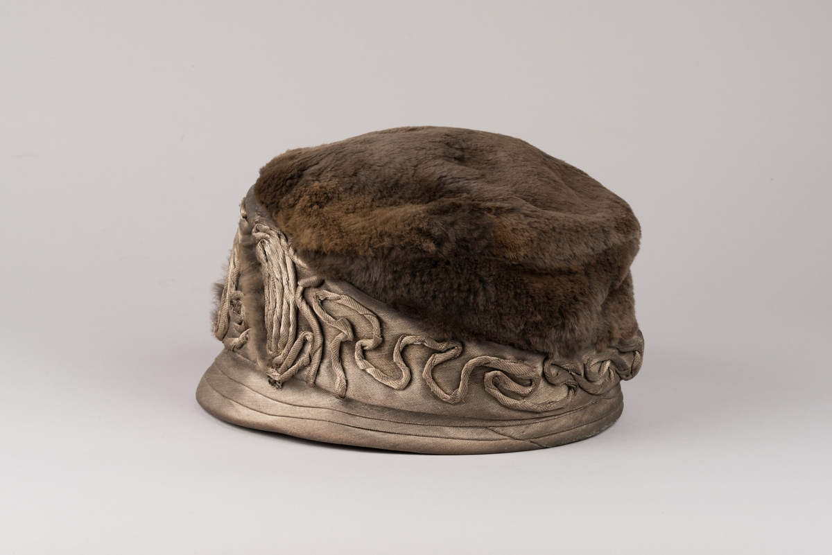 Hatt, modell med brun päls på kullen och grått tyg av metalltråd. Hatten har ett litet styvt brätte och en stärkt kant som är bredare i framkant. Dekorationer av tvinnat tyg och päls.