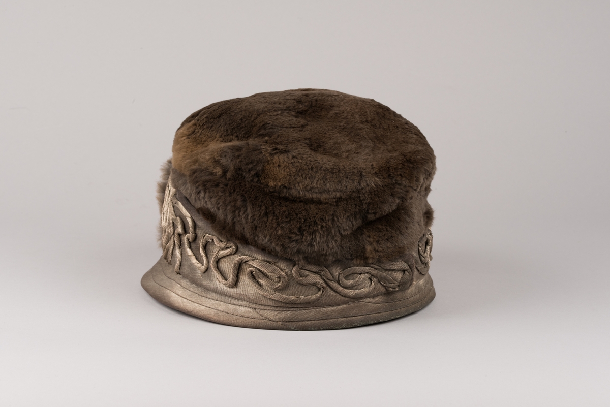 Hatt, modell med brun päls på kullen och grått tyg av metalltråd. Hatten har ett litet styvt brätte och en stärkt kant som är bredare i framkant. Dekorationer av tvinnat tyg och päls.