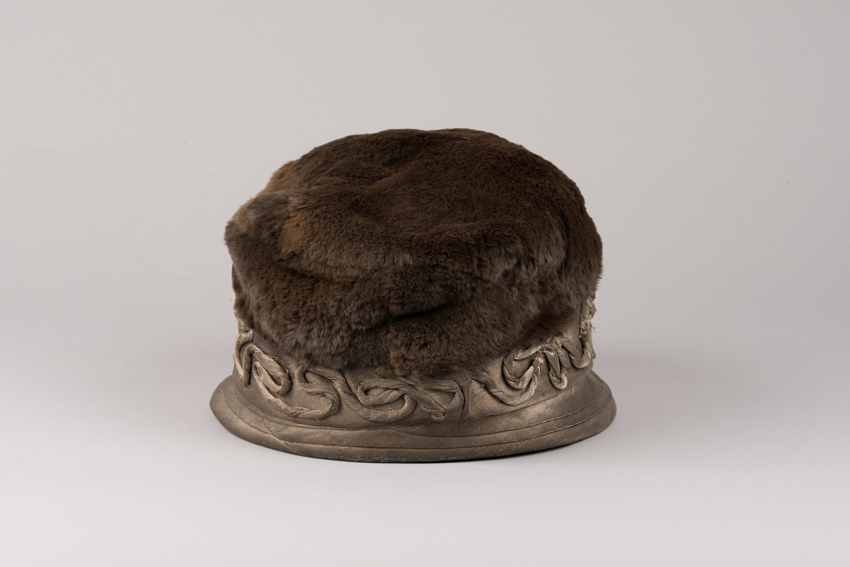 Hatt, modell med brun päls på kullen och grått tyg av metalltråd. Hatten har ett litet styvt brätte och en stärkt kant som är bredare i framkant. Dekorationer av tvinnat tyg och päls.