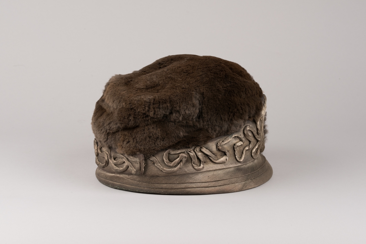 Hatt, modell med brun päls på kullen och grått tyg av metalltråd. Hatten har ett litet styvt brätte och en stärkt kant som är bredare i framkant. Dekorationer av tvinnat tyg och päls.