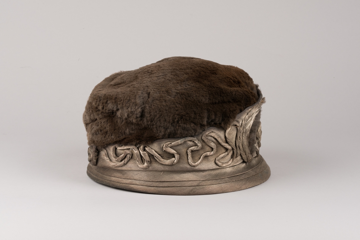 Hatt, modell med brun päls på kullen och grått tyg av metalltråd. Hatten har ett litet styvt brätte och en stärkt kant som är bredare i framkant. Dekorationer av tvinnat tyg och päls.