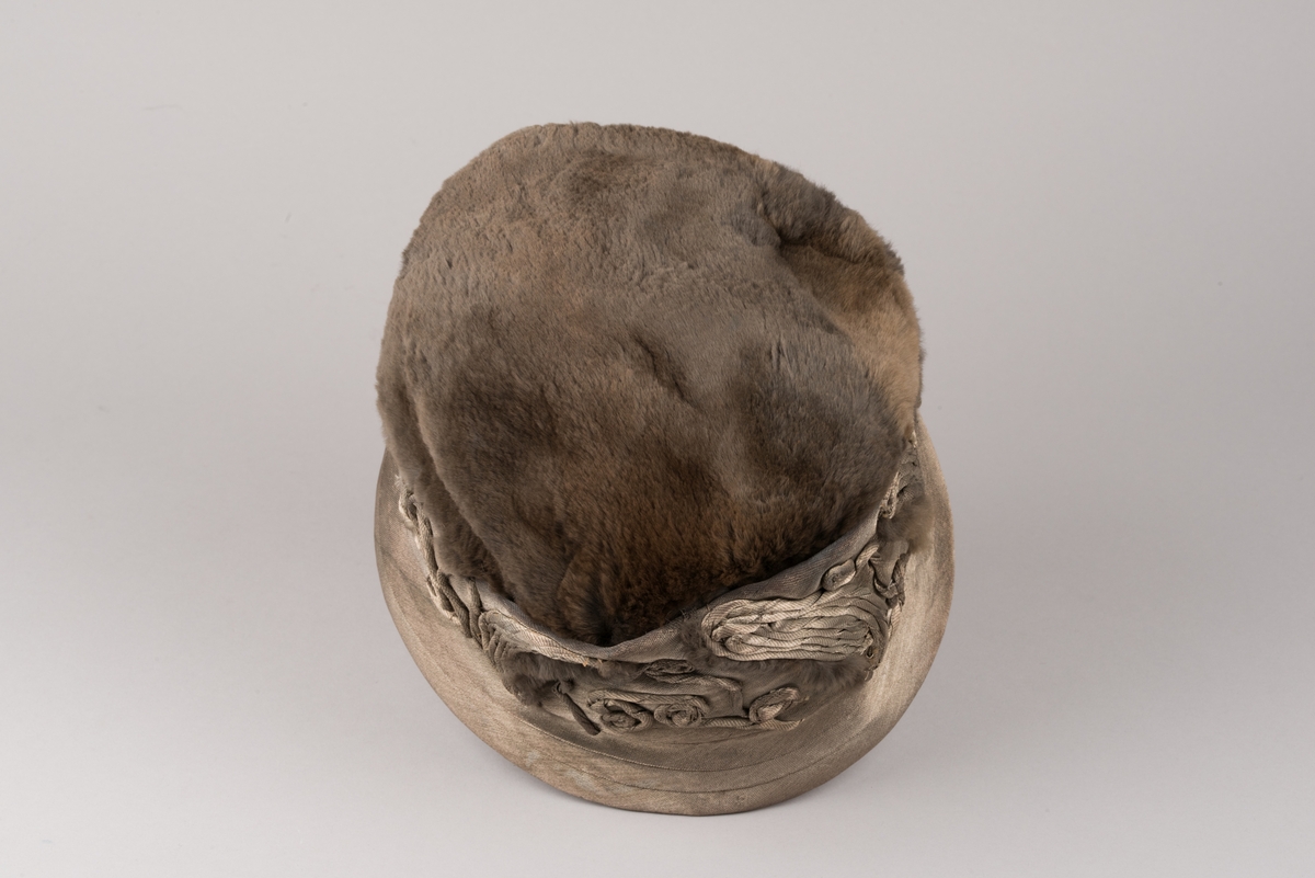Hatt, modell med brun päls på kullen och grått tyg av metalltråd. Hatten har ett litet styvt brätte och en stärkt kant som är bredare i framkant. Dekorationer av tvinnat tyg och päls.