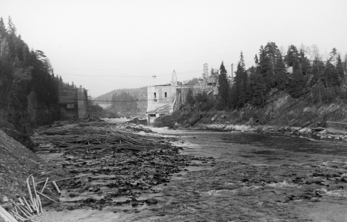 Fra bygginga av den nye kraftverksdammen ved Kykkelsrud i Nedre Glomma, som her var grense mellom Askim og Spydeberg kommuner. Anleggsarbeidet pågikk i perioden 1939-1941. Dette fotografiet skal være tatt høsten 1940, cirka et halvt år før dammen ble ferdig. Bildet er tatt i motstrøms retning, i en periode da vannføringa i elva var svært liten. En del fløtingsvirke lå igjen på ei steinør til venstre i forgrunnen. Bakenfor ser vi hvordan anleggsarbeiderne hadde reist betongkonstruksjoner fra begge elvebredder, men sjølve elveløpet var fortsatt åpent. På tvers av elveløpet skimter vi et par vaierstrekk, som antakelig ble brukt til å transportere materiell til byggeprosessen på taubane. På østsida av elva – til høyre – ser vi et par tårn, som antakelig ble brukt under anleggsarbeidet. Den nye dammen ved Kykkelsrud ble en 120 meter lang og opptil 24 meter høy platedam, utført i armert betong. Den fikk tre flomløp med 17 meter lange og 7,5 meter høye valser og ei sektorluke som var 16 meter bred. Via disse lukene skulle det være mulig å avlede 4 000 kubikkmeter vann i sekundet. Dammen ble bygd slik at det ved seinere utvidelser skulle være mulig å øke kapasiteten med ytterligere 200 kubikkmeter i sekundet. For fløtinga ble det bygd ei betongrenne som gikk i tunnel i berget på vestsida av elveløpet. Her skulle fløtingsvirket kunne ledes forbi dammen – 10-12 000 stokker i timen – med et vannforbruk på cirka 25 kubikkmeter i sekundet. Dette fotografiet er tatt ved inntaket til det som skulle blir tømmerrenna.