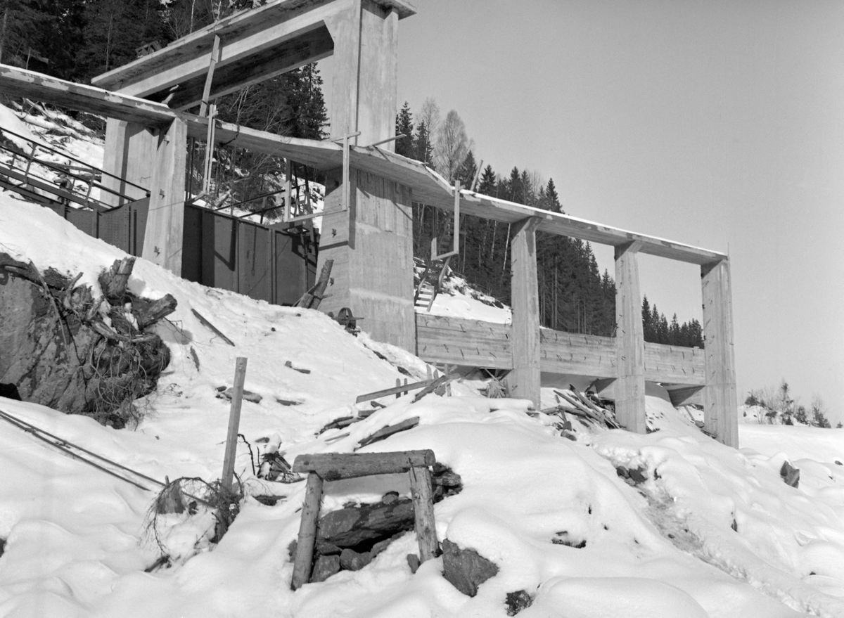 Fra bygginga av dennye kraftverksdammen ved Kykkelsrud i Nedre Glomma, som her var grense mellom Askim og Spydeberg kommuner. Anleggsarbeidet pågikk i perioden 1939-1941. Dette fotografiet skal være tatt den siste anleggsvinteren. Den nye dammen ved Kykkelsrud ble en 120 meter lang og opptil 24 meter høy platedam, utført i armert betong. Den fikk tre flomløp med 17 meter lange og 7,5 meter høye valser og ei sektorluke som var 16 meter bred. Via disse lukene skulle det være mulig å avlede 4 000 kubikkmeter vann i sekundet. Dammen ble bygd slik at det ved seinere utvidelser skulle være mulig å øke kapasiteten med ytterligere 200 kubikkmeter i sekundet. For fløtinga ble det bygd ei betongrenne som gikk i tunnel i berget på vestsida av elveløpet. Her skulle fløtingsvirket kunne ledes forbi dammen – 10-12 000 stokker i timen – med et vannforbruk på cirka 25 kubikkmeter i sekundet. Dette fotografiet er tatt ved inntaket til det som skulle blir tømmerrenna.