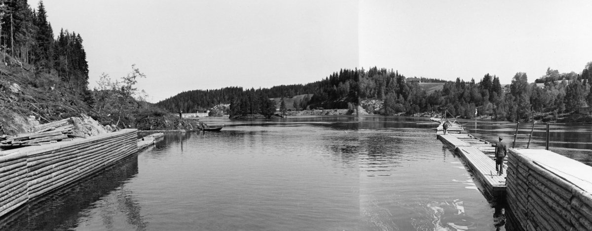 Innløpskanalen til tømmerrenna som ble bygd ved Kykkelsrud kraftstasjon i Nedre Glomma i perioden 1939-1941. Fotografiet er altså tatt i motstrøms retning, oppover vannspeilet, der det (til høyre på dette fotografiet) var lagt ei avstivet ganglense på skrå over vassdraget. Denne lensa skulle tjene både som en skjerm som skulle lede løstømmer som kom flytende ovenfra mot innløpet til tømmerrenna og samtidig tjene som arbeidsplattform for fløterne. Da dette fotografiet ble tatt foregikk det ikke fløting, og de to karene som gikk på lensegangene var antakelig på befaring ved dette nyanlegget. Både til venstre og (så vidt) ved høyre bildekant skimter vi såkalte «skådammer», skjermer av tømmer som også skulle lede tømmer mot renna. En liten robåt lå fortøyd ved en odde til venstre i bildet. Fotografiet er tatt i 1941, som var den første sesongen den nye dammen og tømmerrenna ved Kykkelsrud var i drift.

Mer informasjon om kraftproduksjonen ved Kykkelsrud og det nevnte byggeprosjektet finnes under fanen «Opplysninger».