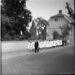 Flickor går två och två på led längst med Brahegatan. Kyrkoherde Wallis Wendel leder dem. Längst bak går några pojkar.