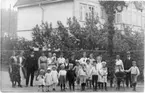 En grupp kvinnor, män och barn står vid en midsommarstång, barnen håller varandras hand. I bakgrunden ett större hus med träpanel och inglasad veranda.