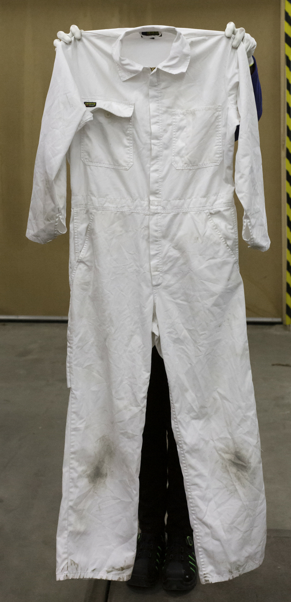 Skyddsoverall i vitt tyg, kypertvävt. Sammansättning 65% polyester, 35% bomull. Storlek C48. Två bröstfickor , två sidfickor och slitsar för att nå innanliggande kläder, två bakfickor samt en avlång benficka på höger ben. Gummiband för justering av midjemått. Tvåvägs dragkedja av metall märkt YKK.
Smutsig på knän, armbågar och bakdel.