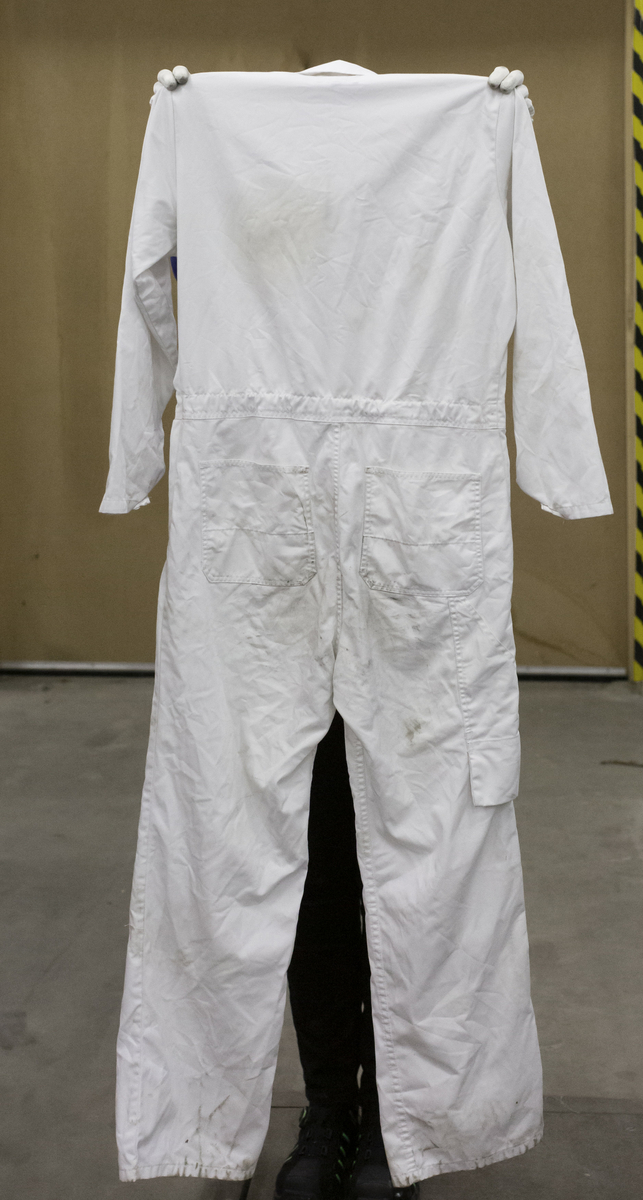 Skyddsoverall i vitt tyg, kypertvävt. Sammansättning 65% polyester, 35% bomull. Storlek C48. Två bröstfickor , två sidfickor och slitsar för att nå innanliggande kläder, två bakfickor samt en avlång benficka på höger ben. Gummiband för justering av midjemått. Tvåvägs dragkedja av metall märkt YKK.
Smutsig på knän, armbågar och bakdel.