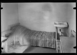 Ett enkelt inrett rum med säng, bord och handfat i hörnet.
F