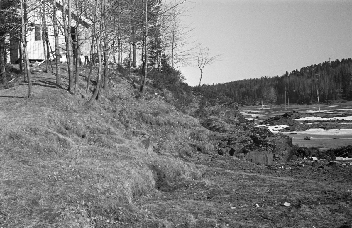 Elvelandskap ved Nedre Glomma. Fotografiet er tatt ved en berglendt elvebredd. På berget øverst til venstre på bildet ligger det som i Glomma fellesfløtingsforenings gamle fotoalbum er kalt «hvilehytten» på «Tjueholmen ovf. Jutulbroen». Denne hytta var antakelig et sted som tømmerfløterne kunne bruke som spise- og kvilebrakke når de arbeidet med å løsne tømmer som hadde satt seg fast på den nevnte Jutulbrua, ei rekke av steinskjær som sperret mye av elveløpet i lavvannsperioder.