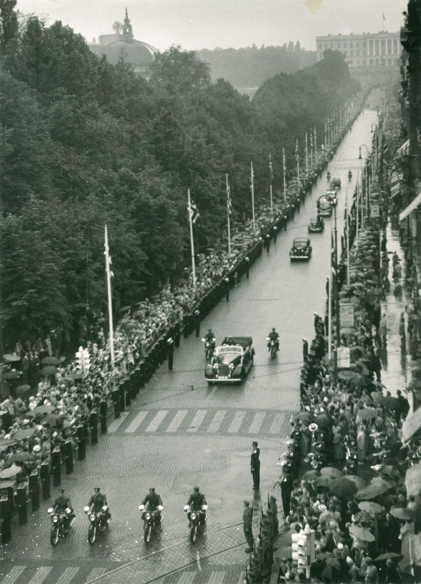 Kong Haakon VII sitter i en åpen bil eskortert av politi på motorsykler på vei nedover Karl Johans gate. Slottet i bakgrunnen.