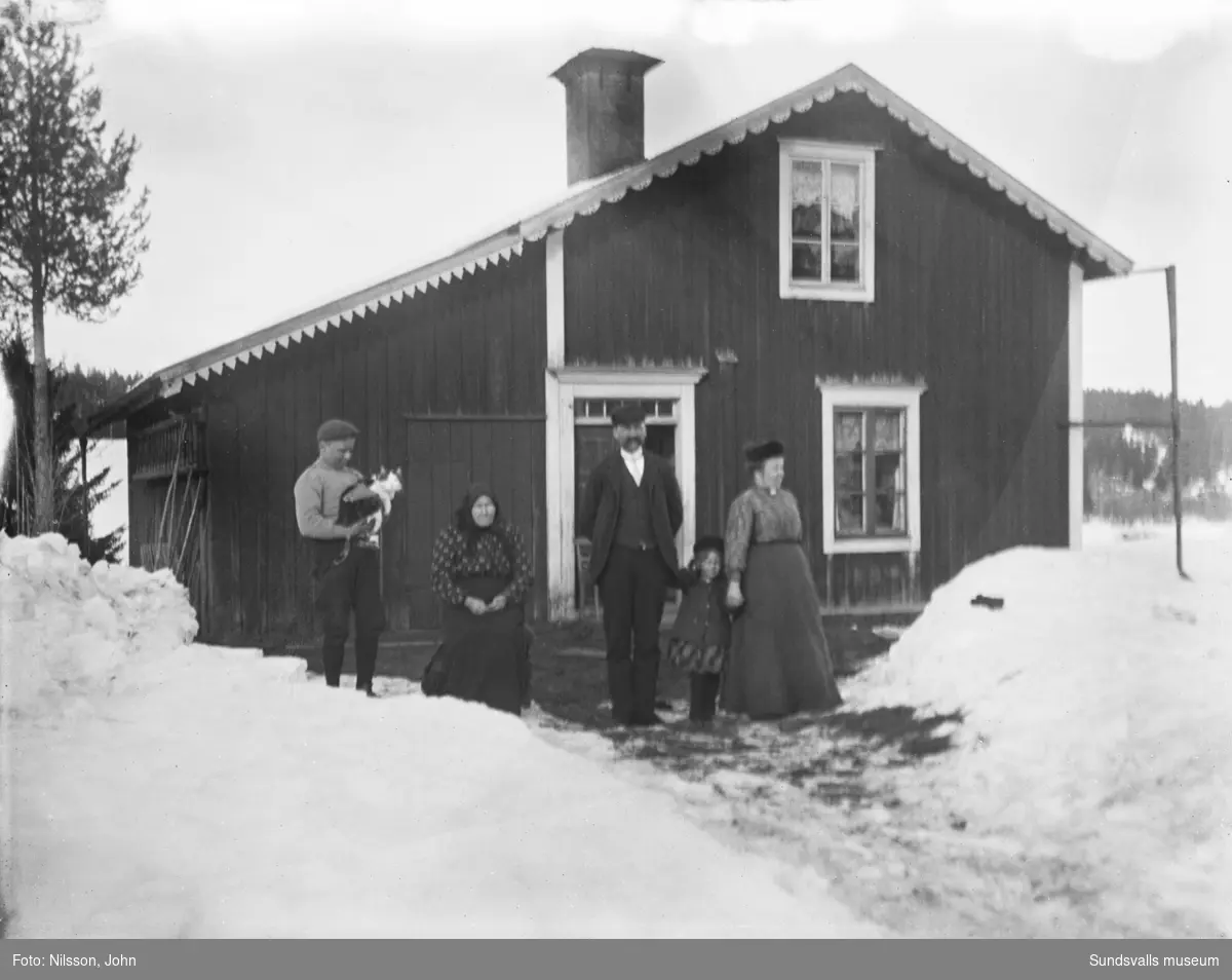 Två vinterbilder ur olika vinklar mot ett mindre bostadshus, framför står två män, två kvinnor, en liten flicka och en katt.