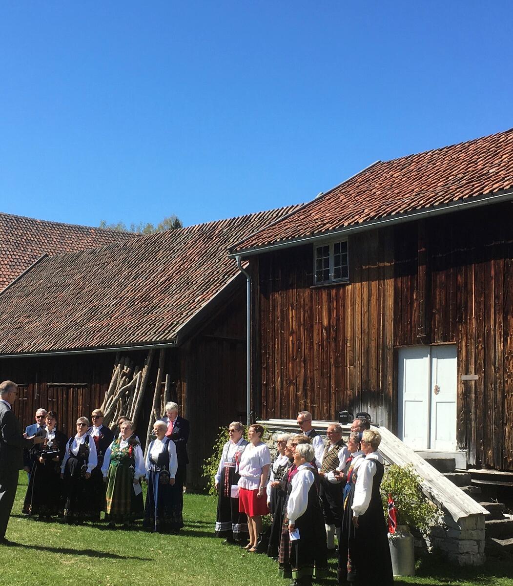 17. mai og kor i bunad foran stabburet på Stenberg, Vestre Toten. 
Blå himmel og sol.