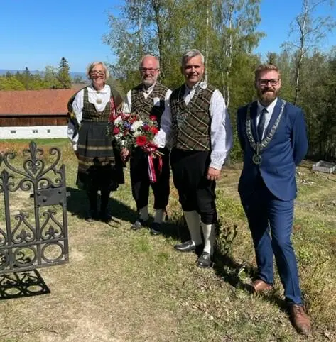 Fire personer, festpyntede representanter for Toten historielag og ordførere i Østre og Vestre Toten står ved gravstedet til Weidemannfamilien, med en blomsterkrans som skal legges på graven. Sol og blå himmel. Låven på Stenberg i bakgrunnen.